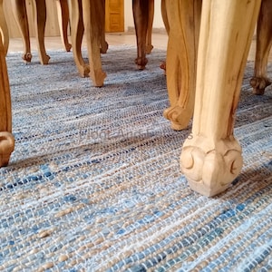 Puede incluir: Primer plano de las patas de madera tallada de una mesa, con un acabado de madera natural. Las patas de la mesa están sobre una alfombra tejida azul y blanca.