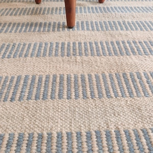 May include: Close-up of a woven rug with alternating stripes of light blue and beige. The rug has a textured appearance and is partially obscured by the wooden legs of a piece of furniture.