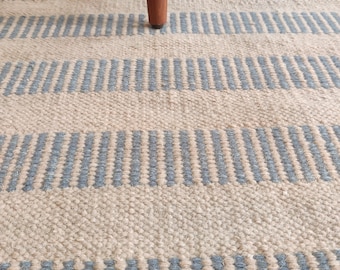 Könnte beinhalten: Nahaufnahme eines gewebten Teppichs mit abwechselnden Streifen in Hellblau und Beige. Der Teppich hat eine strukturierte Oberfläche und wird teilweise von den Holzbeinen eines Möbelstücks verdeckt.