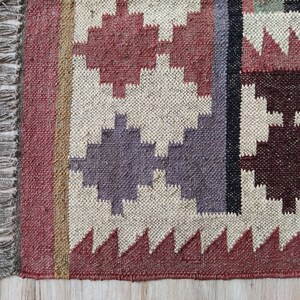 May include: A woven rug with a geometric pattern in shades of red, brown, beige, and gray. The rug has a fringe border.