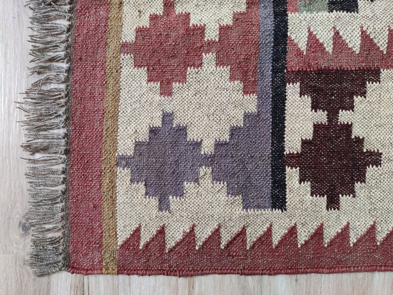 May include: A woven rug with a geometric pattern in shades of red, brown, beige, and gray. The rug has a fringe border.