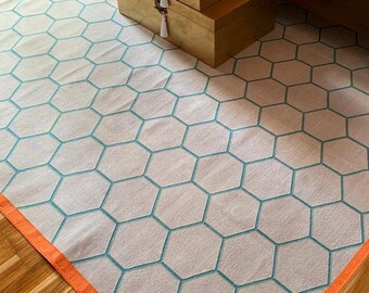 May include: A white rug with a teal honeycomb pattern and an orange border. The rug is laid on a wooden floor.