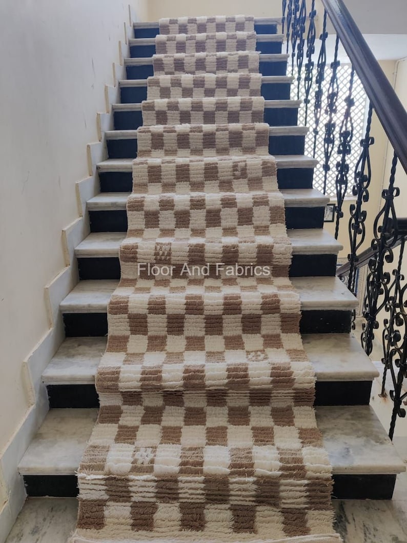 May include: A brown and white checkered rug is laid out on a set of stairs. The rug is made of a soft, fluffy material and has a subtle sheen. The stairs are made of marble and have a black trim. The text "Floor And Fabrics" is visible on the rug.