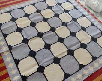 May include: A gray and white checkered rug with a black border. The rug has a geometric pattern and is made of cotton.