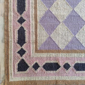 Könnte beinhalten: Ein gewebter Teppich mit geometrischem Design. Der Teppich hat einen Rand mit einem rosa und schwarzen Muster, einen braunen Rand und ein zentrales Rautenmuster in Lavendel und Beige. Der Teppich hat einen Fransenrand.