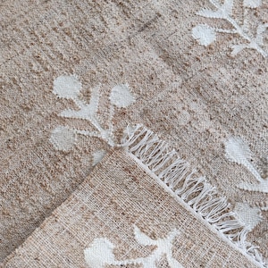 May include: A beige and white woven rug with a floral pattern. The rug has a fringe border and is folded in half.
