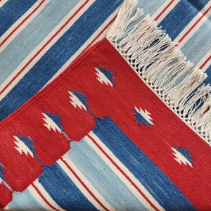 May include: Red, blue, and white striped rug with a fringe border. The rug has a geometric pattern in the center.