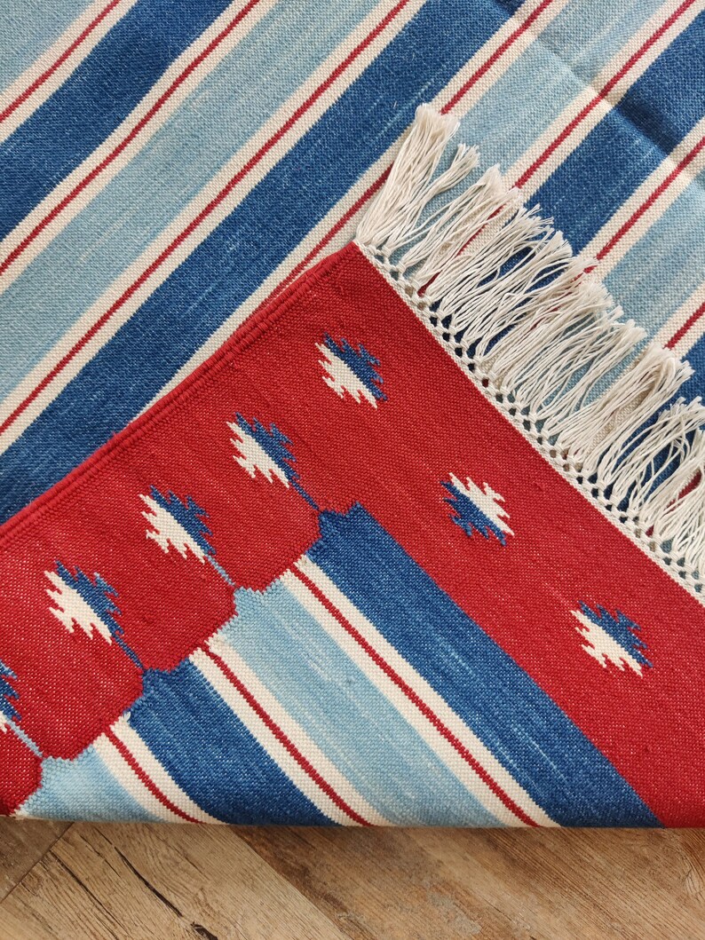 May include: Red, blue, and white striped rug with a fringe border. The rug has a geometric pattern in the center.