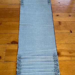 May include: A light blue woven rug with fringed edges. The rug is laid out on a wooden floor.
