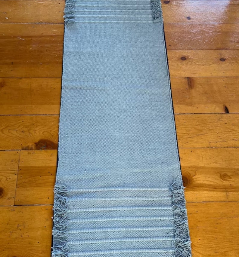 May include: A light blue woven rug with fringed edges. The rug is laid out on a wooden floor.