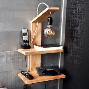 Könnte beinhalten: Ein Wandregal aus Holz mit zwei Ablagen, einer Leuchte mit einer hängenden Glühbirne, einem Telefon, einem Buch und einem kleinen elektronischen Gerät. Das Regal ist an einer grauen Wand montiert.