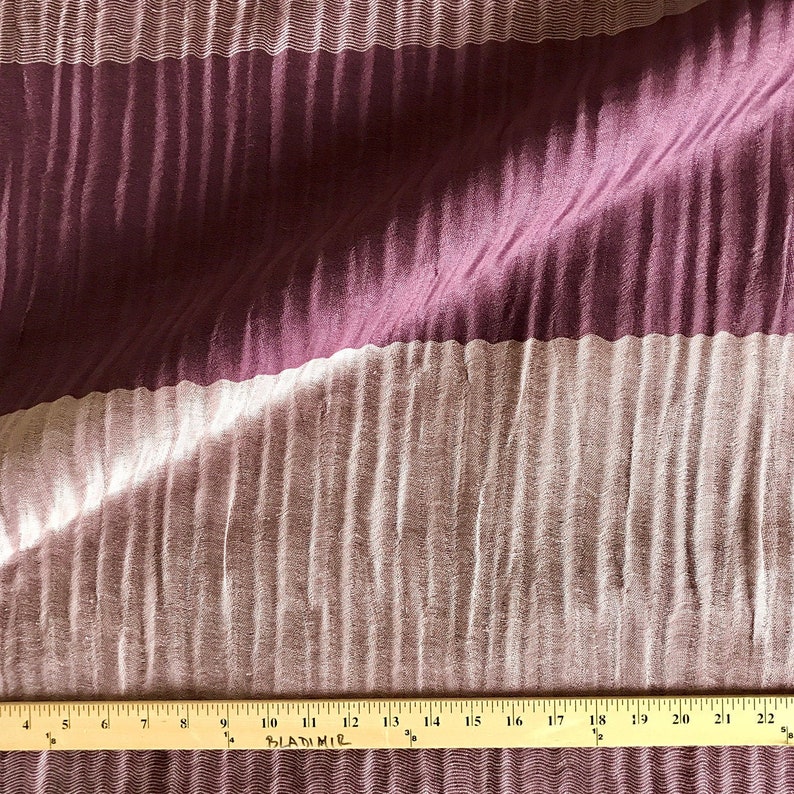 May include: A close-up of a fabric with a striped pattern. The fabric is a combination of a deep purple and a light beige. The fabric is pleated and has a subtle sheen.