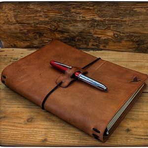 Op de afbeelding: Een bruin lederen notitieboek met een pen die is vastgemaakt met een zwart elastiek en een rode leren band. Het notitieboek heeft een klein vogellogo op de omslag.