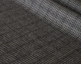 Puede incluir: Una tela textil con un patrón de pata de gallo en gris oscuro y negro. El material tiene una textura tejida visible con un aspecto suave y ligeramente difuso, mostrando pliegues que crean profundidad y sombras en la superficie de la tela.