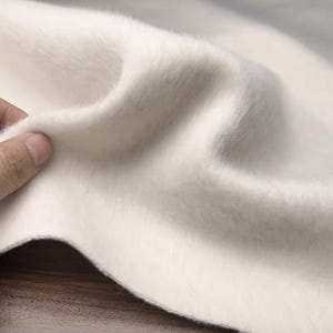 May include: Close-up of a soft, off-white fabric with a fluffy texture. The material is being held, showing its flexibility and drape. The fabric appears to be a plush textile, possibly wool or a similar material.