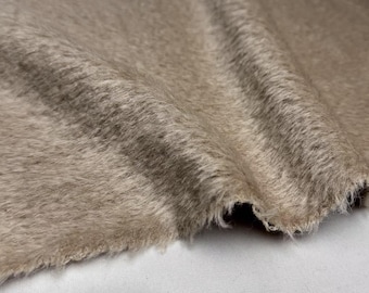 May include: Close-up of a piece of taupe-colored faux fur fabric. The fabric has a soft, textured appearance with long, flowing fibers. The edges of the fabric are frayed, revealing the underlying material. The fabric is draped, showing its flexibility.