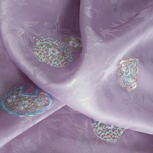 Può includere: Primo piano di un tessuto lavanda con disegni floreali e astratti ricamati. Il tessuto ha una lucentezza sottile ed è drappeggiato, mostrando gli intricati dettagli del ricamo. Il ricamo presenta sfumature di blu, verde e oro.