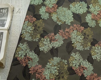 May include: A fabric with a floral pattern featuring clusters of hydrangea blooms in shades of pale green, peach, and muted gold against a dark olive-green background. Dark leaves are interspersed among the flowers.
