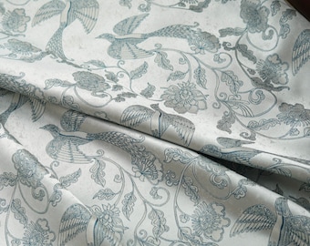 May include: A light-colored fabric featuring a repeating pattern of dark blue birds and intricate floral motifs. The design includes long-tailed birds in flight and various blossoms connected by swirling vines, printed on a smooth, off-white textile.