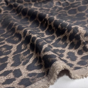 May include: Close-up of fabric featuring a dark grey background with a brown leopard print pattern. The fabric is folded, displaying its texture and design. The edges are finished with a fringed trim.