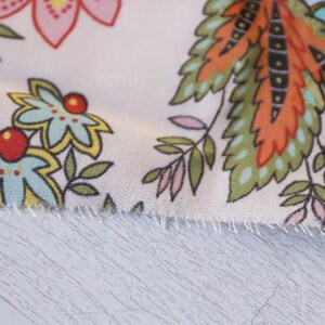 May include: Close-up of a floral fabric with a white background. The fabric features colorful flowers and leaves in shades of pink, blue, yellow, orange, and green. The edges of the fabric are frayed.