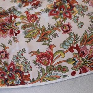 May include: Close-up of a fabric with a floral pattern. The design features large, stylized flowers and leaves in shades of red, orange, green, and blue on a cream background. The fabric has a soft, slightly textured appearance.