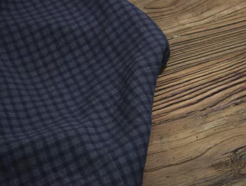 May include: A close-up of a navy blue fabric with a small checkered pattern. The fabric is draped over a wooden surface with visible grain. The fabric's texture and pattern are clearly visible.