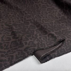 May include: Close-up of a dark brown fabric featuring a leopard print design. The material has a textured feel with frayed edges. The leopard spots are a darker shade of brown, creating a contrast.