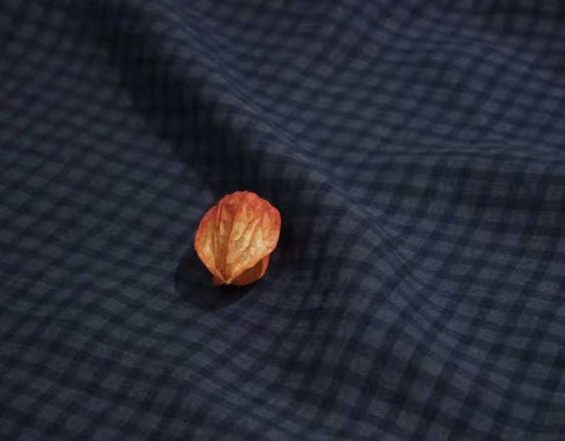 May include: A close-up shot of a piece of navy blue gingham fabric with a dried orange-colored seed pod resting on top. The fabric is slightly wrinkled, creating shadows and texture.