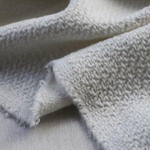 May include: Close-up of a textured, off-white fabric. The material has a looped, nubby surface and a woven edge with frayed threads. The fabric is folded, revealing its thickness and texture. Ideal for upholstery or textile projects.