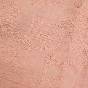 May include: Close-up of a light pink fabric with embroidered floral patterns. The embroidery features detailed, swirling designs of flowers and leaves, creating a delicate, textured surface. The fabric has a slightly wrinkled appearance.