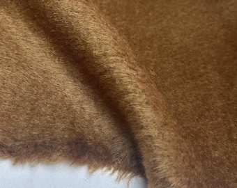 May include: Close-up of a folded piece of brown faux fur fabric. The fabric has a soft, textured appearance with a slightly uneven surface. The color is a warm, earthy brown.