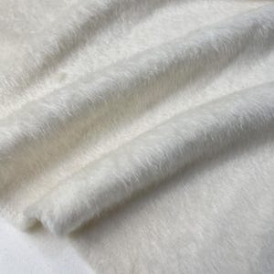 May include: White fluffy fabric with a soft, fuzzy texture. The fabric is folded and draped, showing its texture and drape.