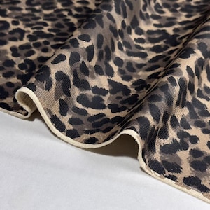 Puede incluir: Primer plano de una tela con estampado de leopardo. La tela presenta un fondo beige con manchas negras y marrones. El borde de la tela está rematado con un ribete color crema. La tela está doblada, mostrando su textura y diseño.