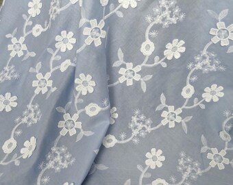 May include: Light blue fabric with a delicate white floral embroidery pattern. The design features detailed white flowers and leaves, creating a flowing, elegant aesthetic. The fabric is draped, showing the texture and depth of the design.