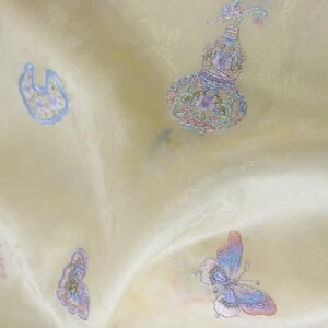 May include: Close-up of a pale yellow fabric with embroidered designs. The fabric features several embroidered motifs, including a butterfly, a gourd-shaped vase, and other floral elements in pastel colours. The fabric has a subtle sheen.