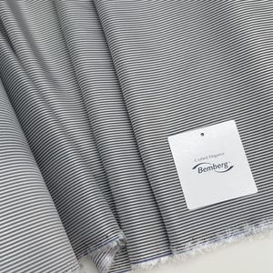 Op de afbeelding: Close-up van een stof met een grijs en wit gestreept patroon. De stof is gevouwen, wat de textuur en de drapering laat zien. Een wit label met de tekst "Crafted Elegance Bemberg" is bevestigd.