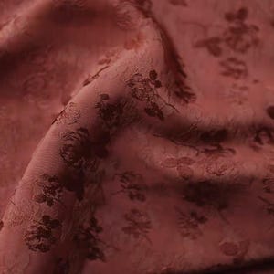 Puede incluir: Primer plano de una tela burdeos con un estampado floral. La tela tiene una superficie texturizada y un brillo sutil. El diseño floral es más oscuro que el color base, creando un contraste. La tela está doblada, mostrando profundidad y dimensión.
