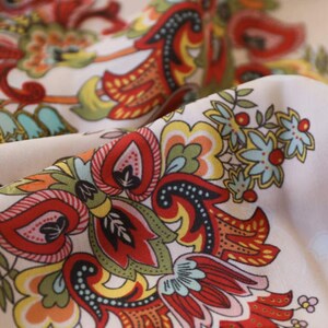 May include: Close-up of a fabric with a detailed floral pattern. The design features red, orange, yellow, green, and blue elements on a cream background. The fabric is folded, showcasing the intricate design and color variations.