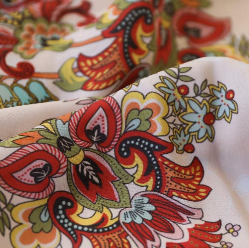 May include: Close-up of a fabric with a detailed floral pattern. The design features red, orange, yellow, green, and blue elements on a cream background. The fabric is folded, showcasing the intricate design and color variations.