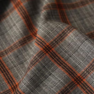Puede incluir: Primer plano de tela gris con un patrón de cuadros. El cuadro presenta líneas naranjas y negras sobre un fondo gris. La tela tiene una apariencia texturizada, con hilos tejidos visibles. La imagen resalta la textura y las variaciones de color del material.