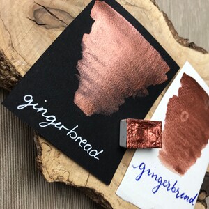 May include: A close-up of a copper-colored watercolor paint pan labeled "gingerbread". The paint is shown on both black and white paper, showcasing its metallic sheen.