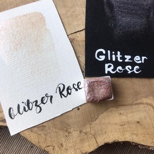 Könnte beinhalten: Eine kleine Dose mit roségoldener Glitzerfarbe, mit einer Probe der Farbe auf einer weißen und einer schwarzen Karte. Der Text "Glitzer Rose" ist auf der schwarzen Karte geschrieben.