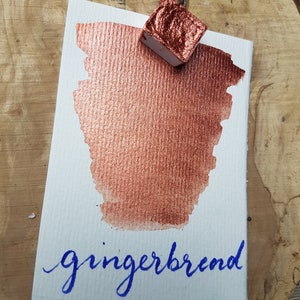 May include: A small pan of metallic copper watercolor paint with the word "gingerbread" written in blue ink on a white paper card.