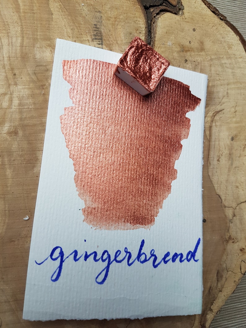 May include: A small pan of metallic copper watercolor paint with the word "gingerbread" written in blue ink on a white paper card.