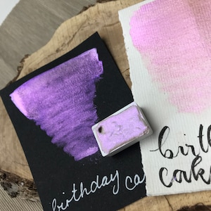 Könnte beinhalten: Eine violette Aquarell-Farbpalette mit dem Text "birthday cake" in weißer Tinte auf einer schwarzen Karte. Die Farbpalette befindet sich neben einem Farbmuster der violetten Farbe auf einer weißen Karte.