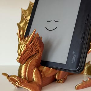 Könnte beinhalten: Ein goldfarbener Drachen-Tablethalter. Der Drache hält ein Tablet mit einem lächelnden Gesicht auf dem Bildschirm. Der Drache hat detaillierte Merkmale und eine strukturierte Oberfläche. Das Tablet ist schwarz mit der Marke "tolino".