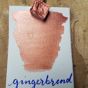 May include: A small pan of metallic copper watercolor paint with a metallic copper paint swatch on a white card with the word "gingerbread" written in blue ink.