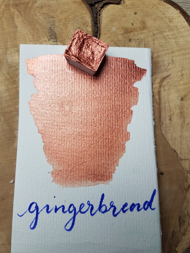 May include: A small pan of metallic copper watercolor paint with a metallic copper paint swatch on a white card with the word "gingerbread" written in blue ink.