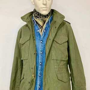 May include: A green military-style jacket with a zipper closure and multiple pockets. The jacket is worn over a blue denim shirt and a patterned scarf.